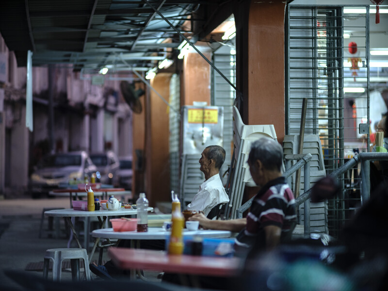 Through a storefront window, diners sit at tables in a casual restaurant or food venue with industrial ceiling and warm lighting.