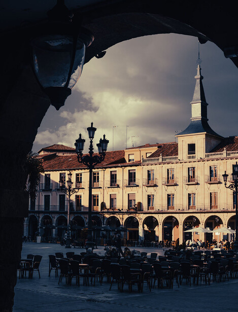 European plaza with classical architecture framed through a dark archway, dramatic cloudy sky illuminating cream-colored buildings.