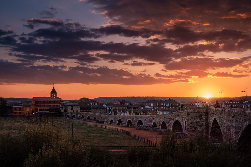 Historic stone bridge with multiple arches spanning a valley at golden hour sunset, with a church spire visible in the distant town skyline.