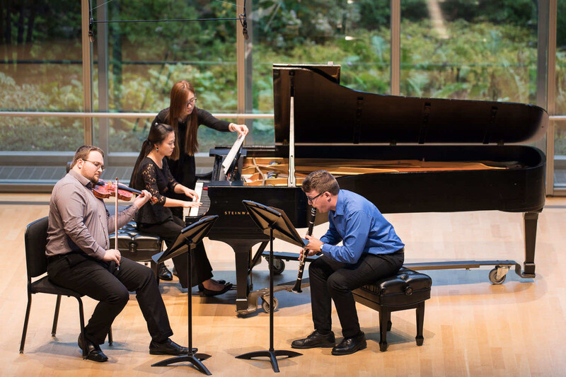 Four musicians perform in a concert hall with a grand piano, music stands, and large windows overlooking trees.
