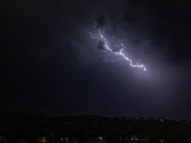 Jagged lightning bolt streaking across a dark stormy sky above a distant hillside silhouette.