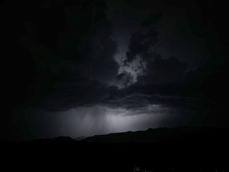 Dramatic storm clouds illuminated by lightning above a dark mountainous horizon at night.