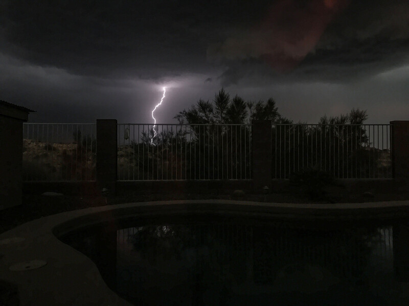 Lightning bolt streaks across dark storm clouds above a residential pool at night.