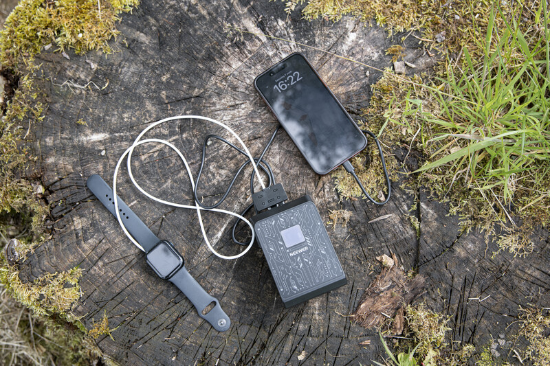 Smartphone, earbuds with white cable, and portable power bank arranged on weathered wood surrounded by moss and grass.