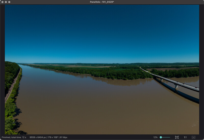 Aerial view of a wide river with muddy brown water cutting through green forest and agricultural land under clear blue sky.