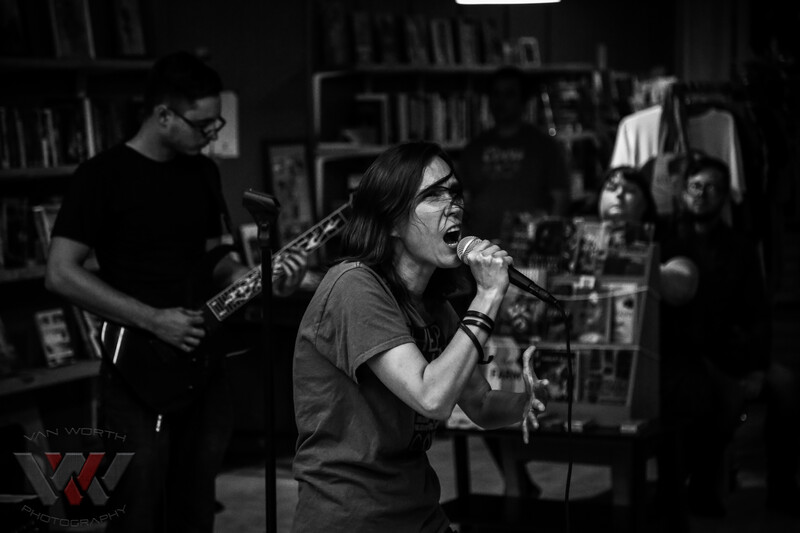 Singer performing at microphone with band members and instruments in dark venue.