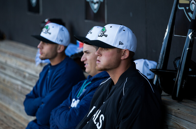 Three baseball players sitting on a bench wearing white caps and team apparel, photographed from the side in natural light.