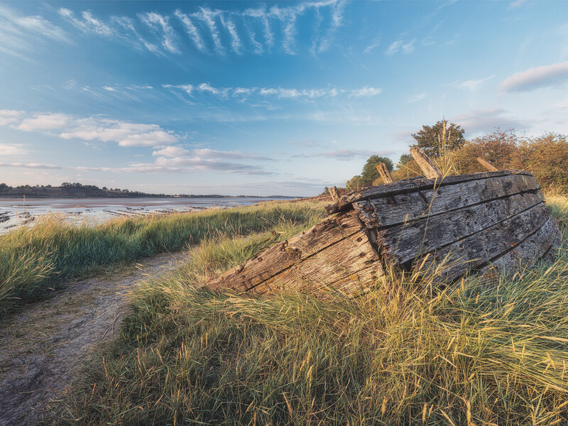 Weathered driftwood log resting on grassy riverbank beside calm water under blue sky.