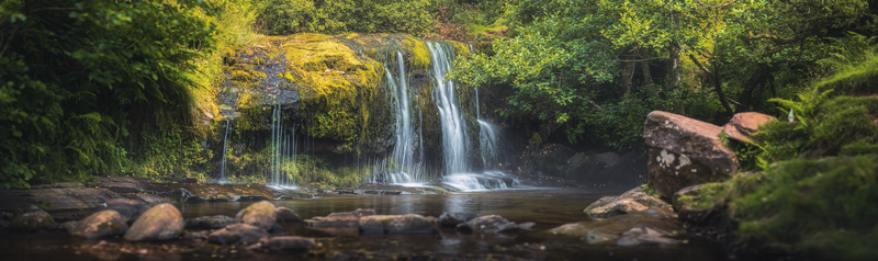 Multi-tiered waterfall cascading over moss-covered rock face surrounded by dense green forest.
