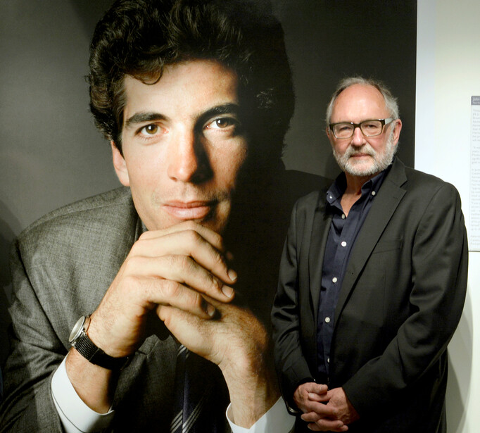Man in dark jacket standing beside large portrait photograph of a young man with dark hair.