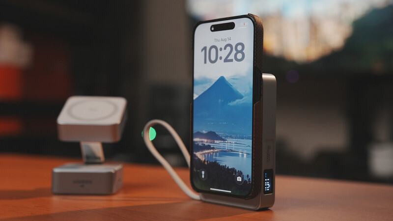 Smartphone displaying a mountain landscape at 10:28 with charging cable attached, positioned on wooden desk next to wireless charging pad.