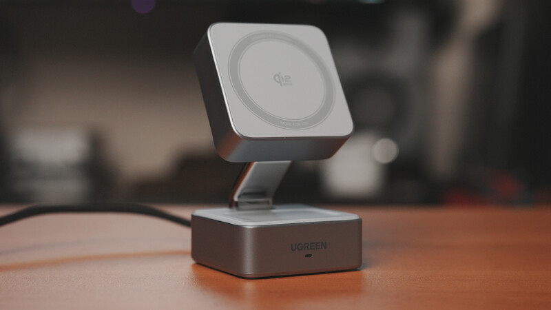 Wireless charging dock with square white pad mounted on dark rectangular base, displayed on wooden desk with blurred background.