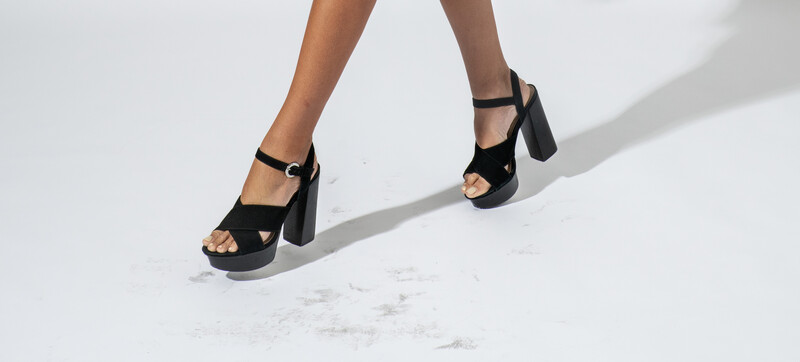 Legs wearing black platform sandals with chunky heels and ankle straps, cast sharp shadows on white marble floor.
