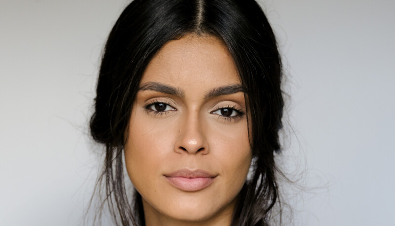 Woman with long dark hair and neutral makeup against a plain gray background.