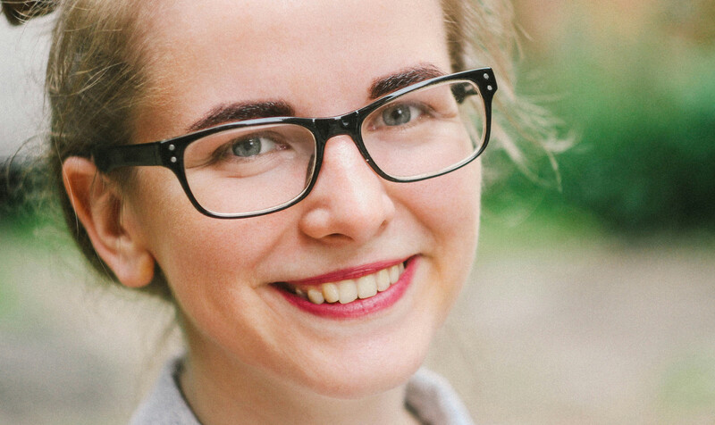 Portrait of a smiling person wearing black-framed glasses and pink lipstick against a soft, blurred green background.