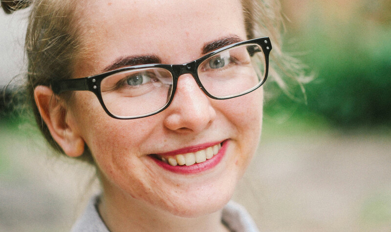 Woman wearing black-framed glasses and pink lipstick, smiling at camera outdoors.
