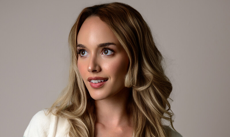 Woman with long wavy blonde hair and pink lipstick against neutral gray background.