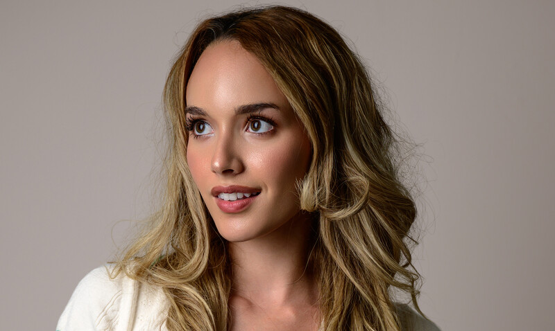 Portrait of a woman with wavy blonde-brown hair and pink lipstick against a neutral gray background.