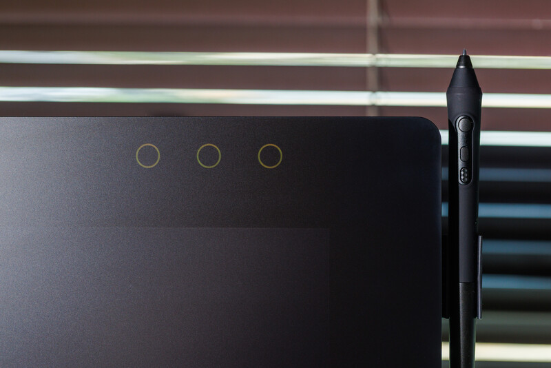 Black digital stylus pen positioned next to a tablet device with three circular indicator lights visible on its dark surface.