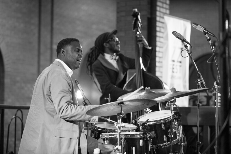 Drummer in light-colored suit playing drums during a jazz performance in a studio setting.