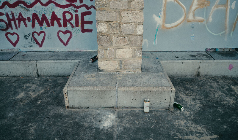 Urban street scene with graffiti-covered wall, concrete pillar, and scattered debris on pavement.
