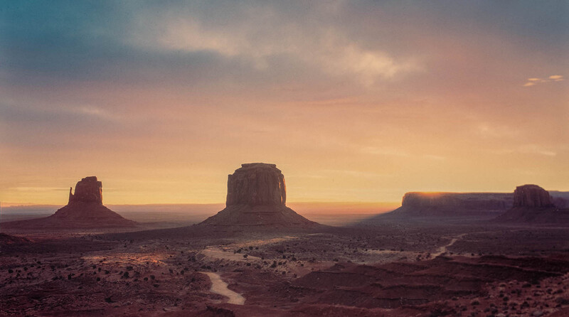 Three massive buttes rising from a desert plain at sunrise, with warm golden and pink light illuminating the sky and rock formations.