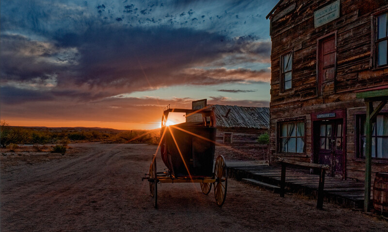 Wooden wagon silhouetted against dramatic sunset beside weathered brick building in desert landscape.