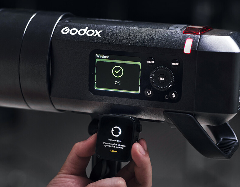 Godox wireless flash trigger held in hand next to a mounted Godox strobe with display screen showing wireless connection status.