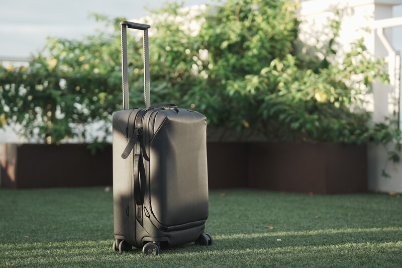 Black rolling suitcase with extended handle standing on grass in a residential backyard setting.
