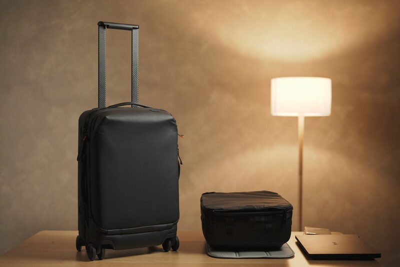 Black rolling suitcase and smaller travel bag arranged on a wooden surface with a lit floor lamp in warm interior lighting.