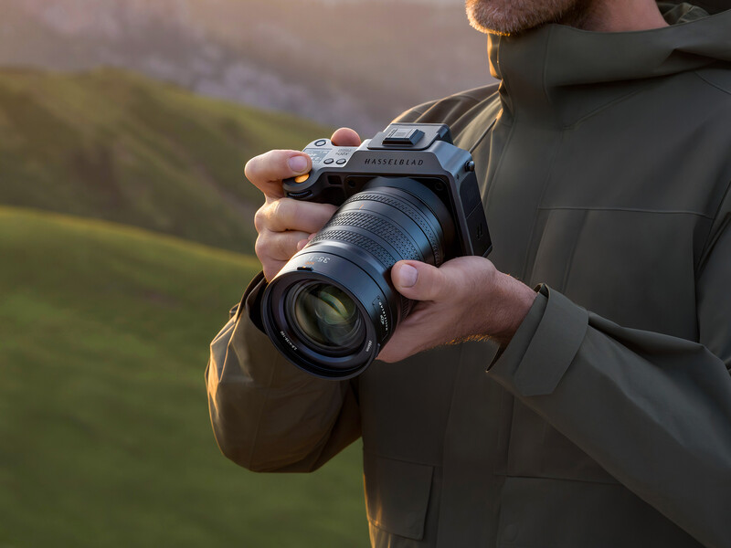 Photographer holding a mirrorless camera with telephoto lens in golden hour light overlooking a blurred green landscape.