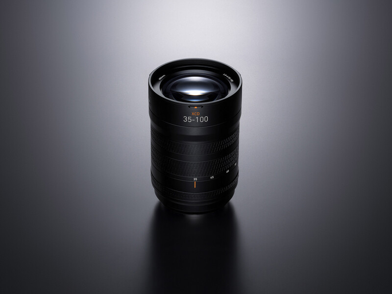 Black zoom lens with 35-100mm focal length marking displayed against neutral gray background.