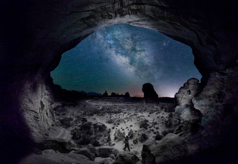 Silhouetted figure standing in rocky cave opening framing a starry night sky and Milky Way above desert landscape.