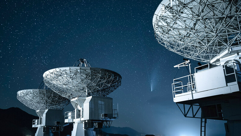 Radio telescope dishes silhouetted against starry night sky with beam of light.
