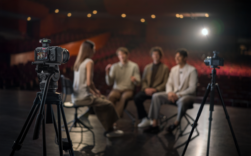 Professional video cameras on tripods recording a panel discussion with five people seated on stage.
