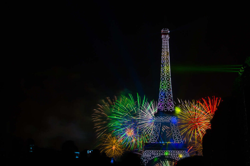 Illuminated Eiffel Tower replica surrounded by colorful fireworks bursting against a dark night sky.