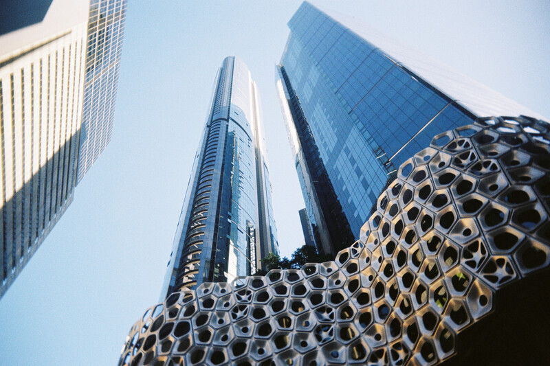Upward view of modern skyscrapers with geometric metal facade in foreground against clear blue sky.