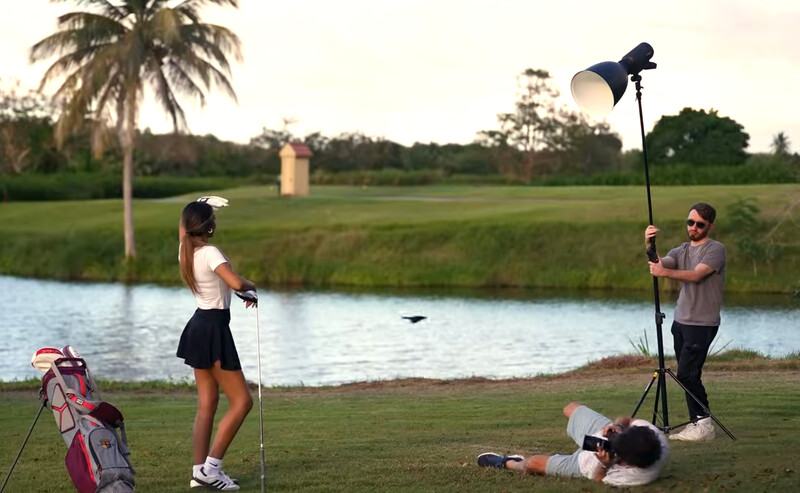 Photographer holding a reflector on a tripod while shooting a female golfer on a grass course near water.