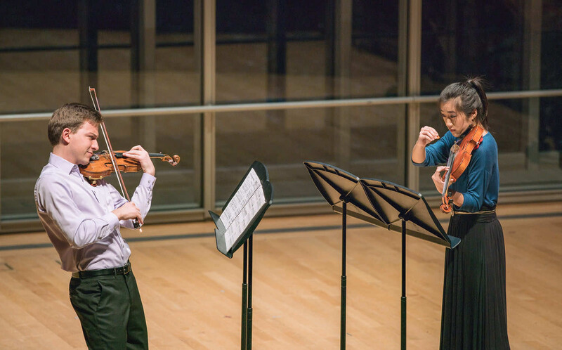 Two violinists perform together in a concert hall, reading sheet music from stands.