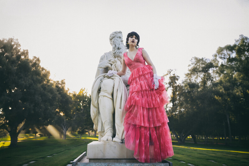 Woman in pink tiered gown posing beside classical stone statue in park setting.