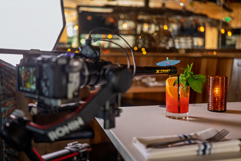 Professional video camera on rig filming colorful cocktails on a white surface with bar background.