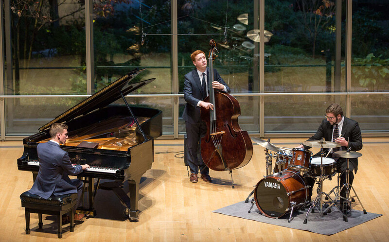Three musicians performing in a concert hall with a pianist, cellist, and drummer on a wooden stage.