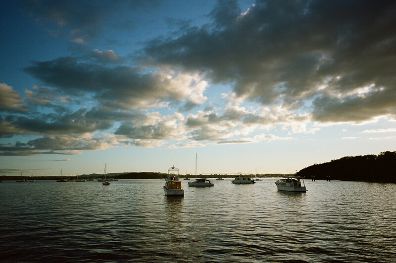 Sailboats anchored in calm water at golden hour beneath dramatic cloud-filled sky.