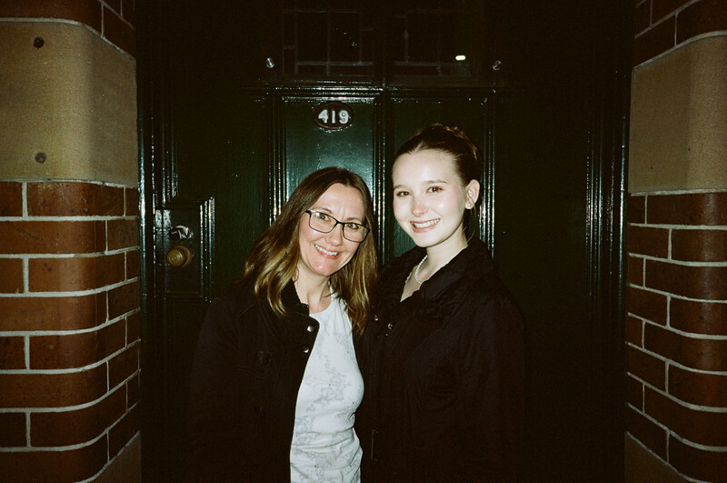 Two women smiling at camera in a hallway lined with brick pillars and dark doors.