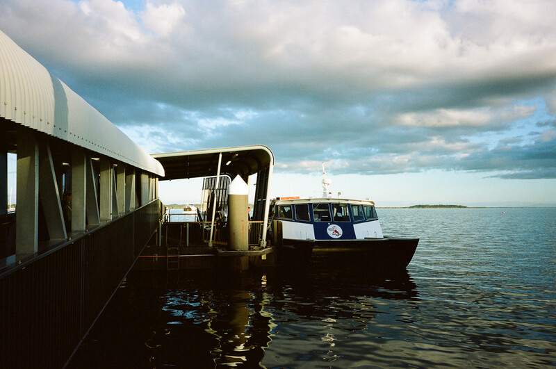 A moored passenger ferry beside a covered dock structure with calm water and dramatic cloudy sky.