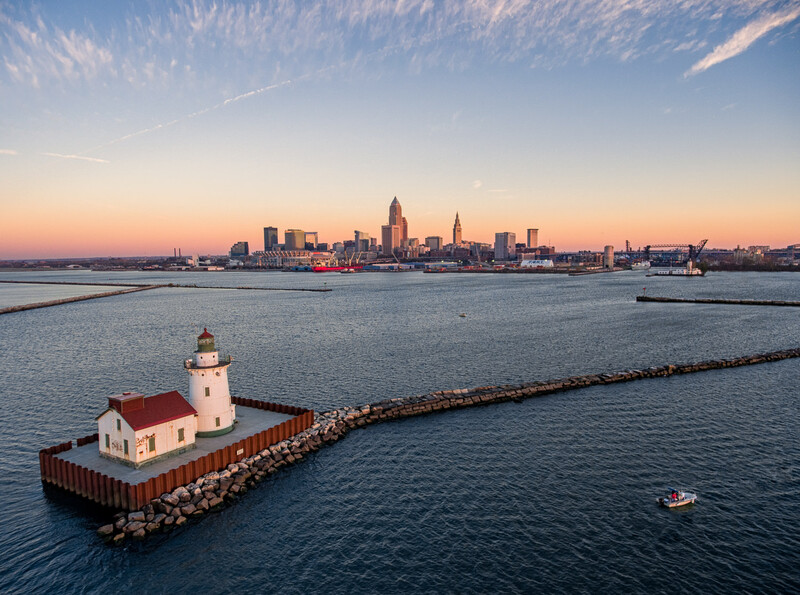 Red and white lighthouse on rocky breakwater at dawn, with city skyline across the water.