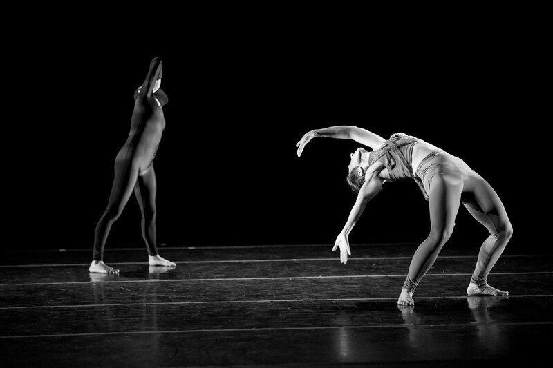 Two dancers in contrasting poses on a dark stage, one upright with arms raised, the other bent forward in deep flexion.