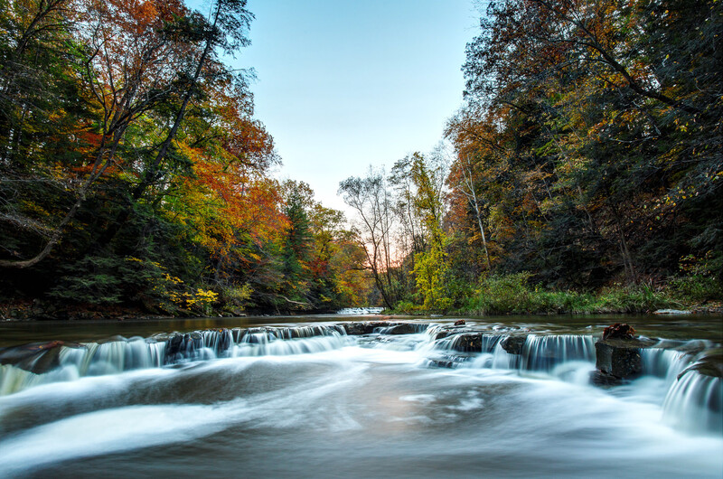 Flowing river with multiple cascading waterfalls surrounded by autumn foliage and dense forest.