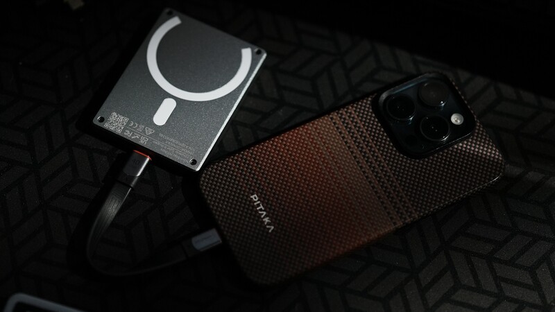 Smartphone with textured case and circular camera setup next to a magnetic charging accessory on dark patterned fabric.