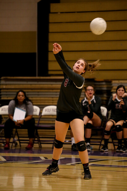 Volleyball player jumping to serve with one arm extended upward, ball visible above her head in a gymnasium.